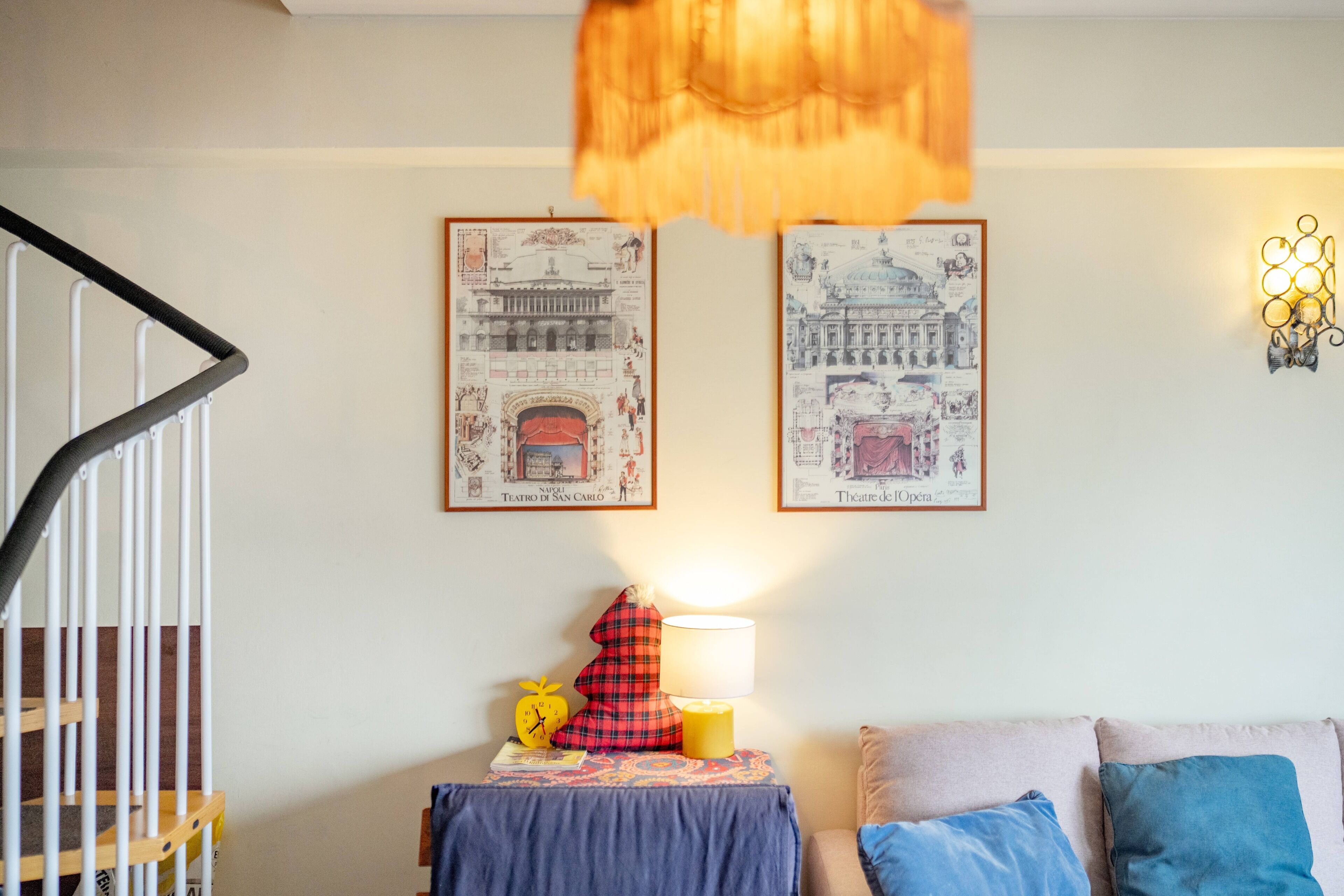 Une pièce avec un escalier, un lit recouvert d’une couverture bleue, une table de chevet avec une lampe, et deux tableaux encadrés accrochés au mur.
