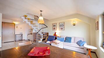 A modern living room with a wooden dining table, a white sofa, a staircase, and a kitchen area.