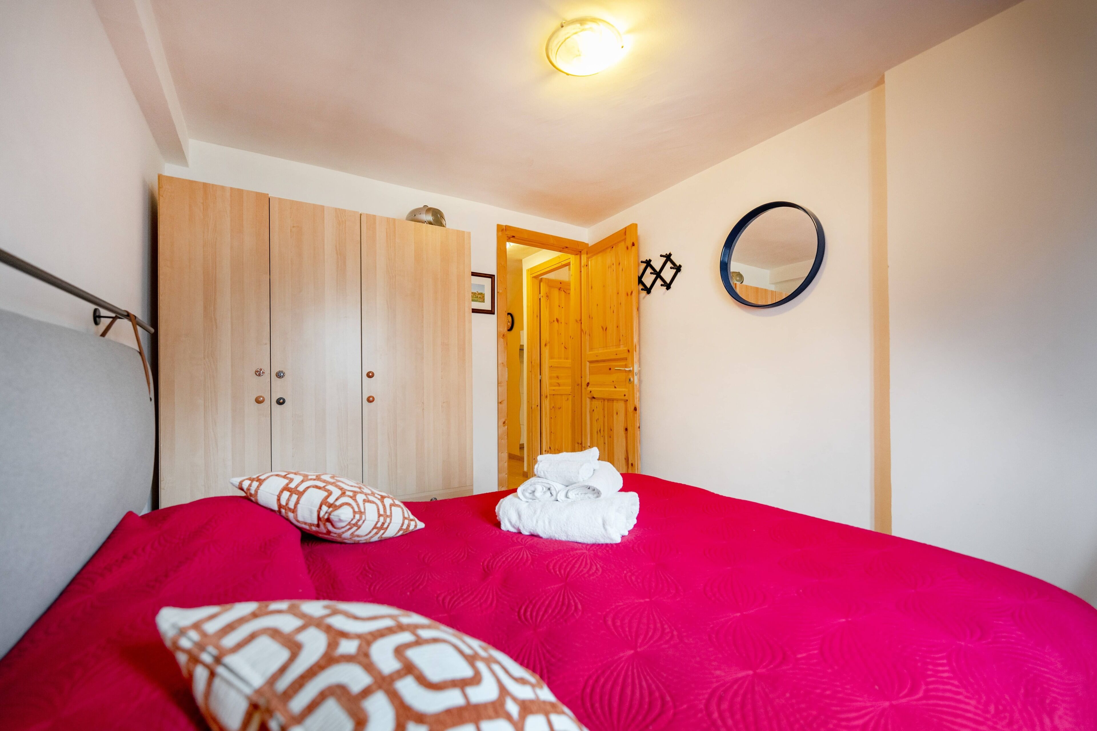 Une chambre à coucher avec un lit, des oreillers, une armoire en bois, un miroir et une porte donnant sur une autre pièce.