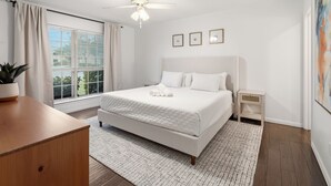 Ein Schlafzimmer mit Bett, Deckenventilator, Fenster mit Vorhängen, Nachttisch und einem Wandbild.