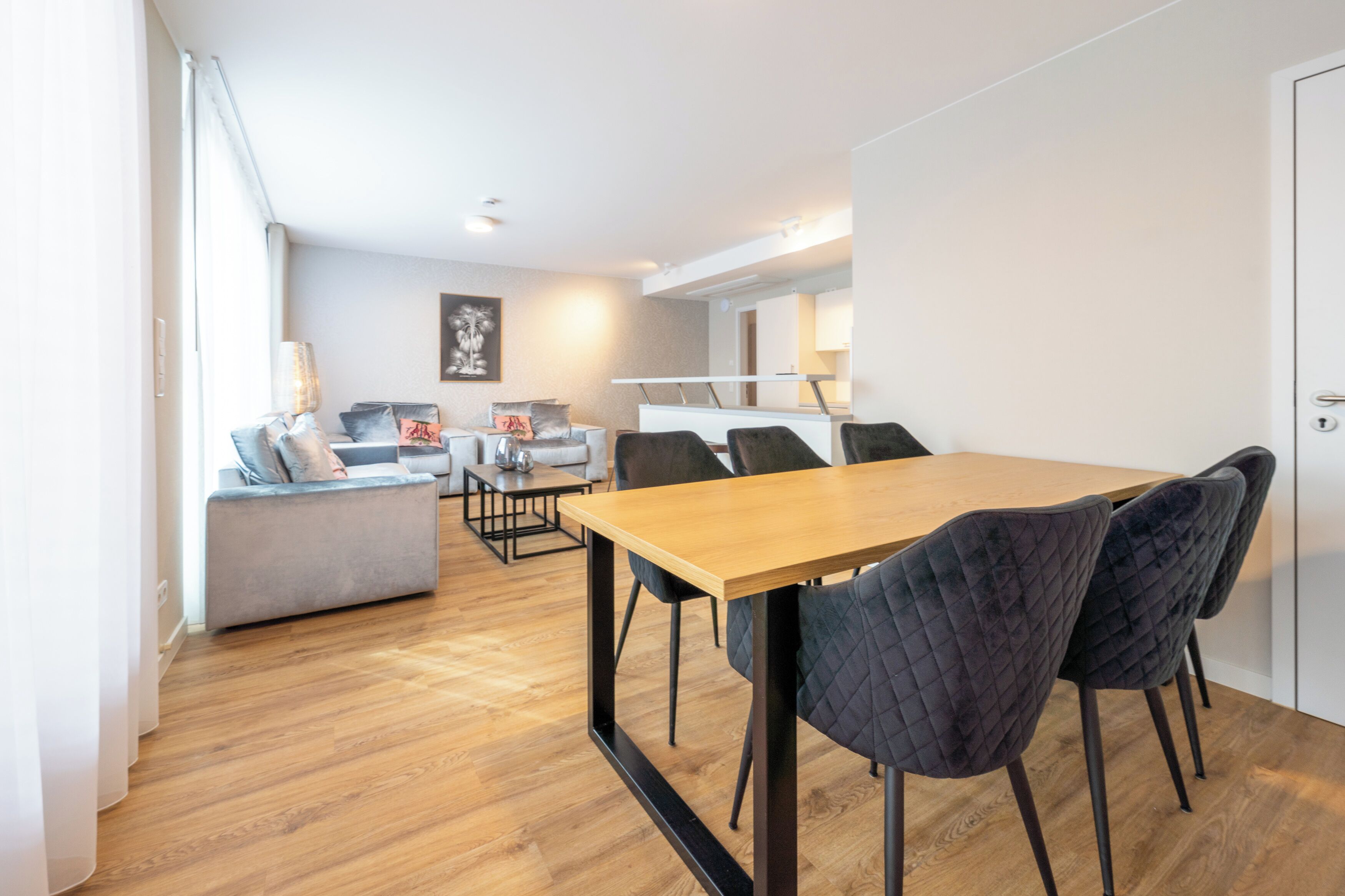 Een moderne woonkamer met een houten eettafel, donkere gestoffeerde stoelen, een grijze bank en een klein bijzettafeltje.