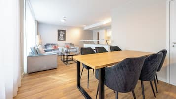 Een moderne woonkamer met een houten eettafel, donkere gestoffeerde stoelen, een grijze bank en een klein bijzettafeltje.