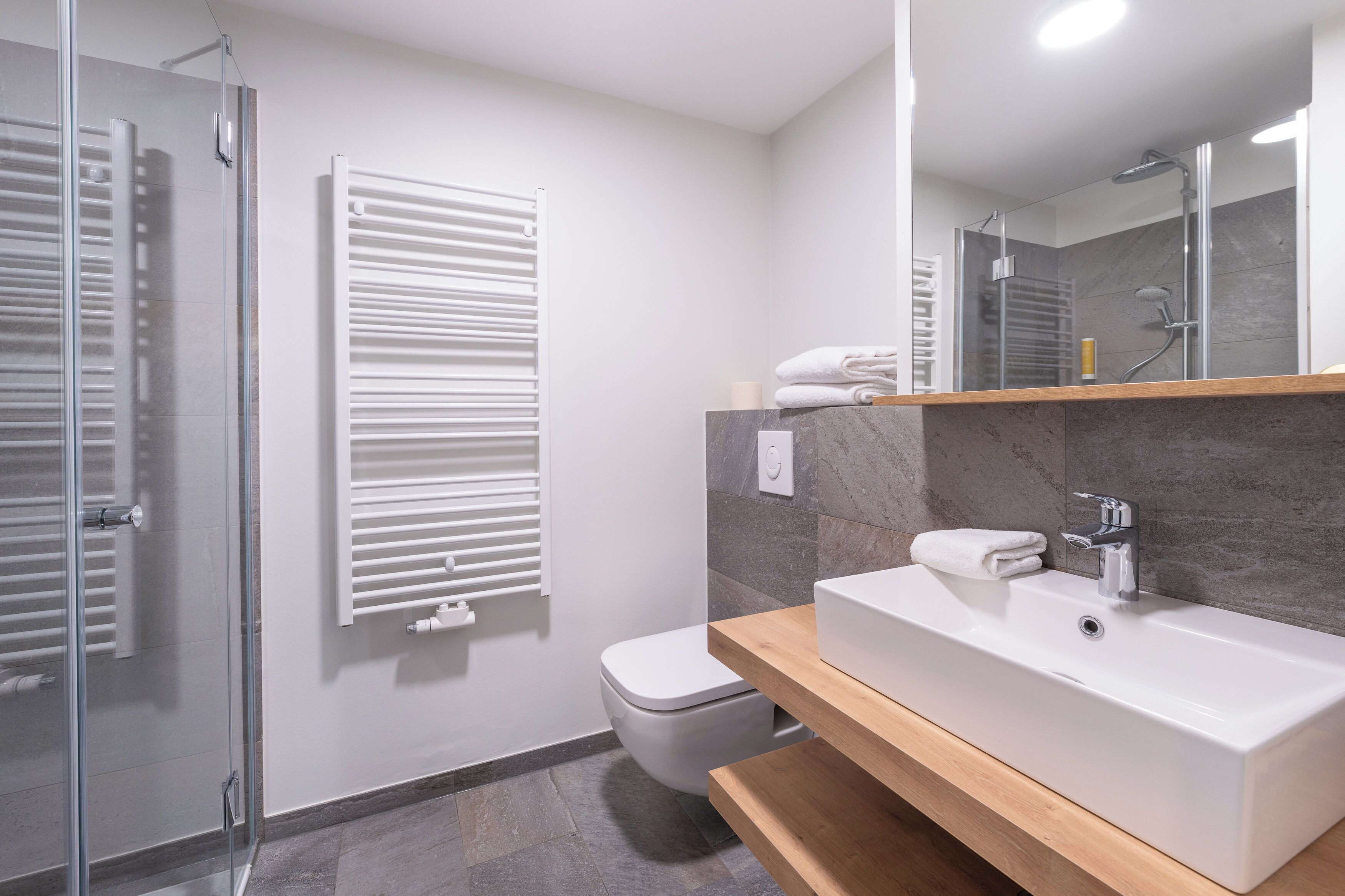 Et moderne badeværelse med en glasbrusekabine, et hvidt toilet, en hvid vask med marmorbordplade og en håndklæderadiator med foldede håndklæder.