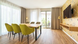 A dining area with a wooden table and green chairs, a TV on the wall, and a balcony with curtains.