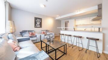 A modern living room with a grey sofa set, a coffee table, and a kitchen area with white cabinets and bar stools.
