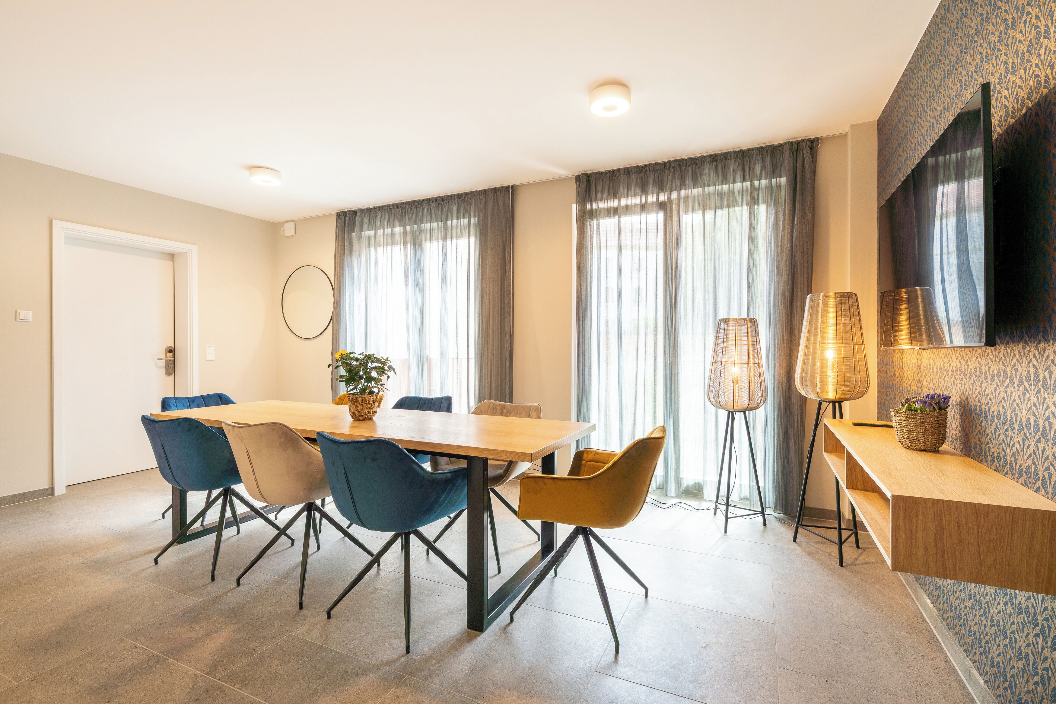 Ein moderner Essbereich mit Holztisch, blau-gelben Stühlen, einer Topfpflanze und einer Stehlampe.