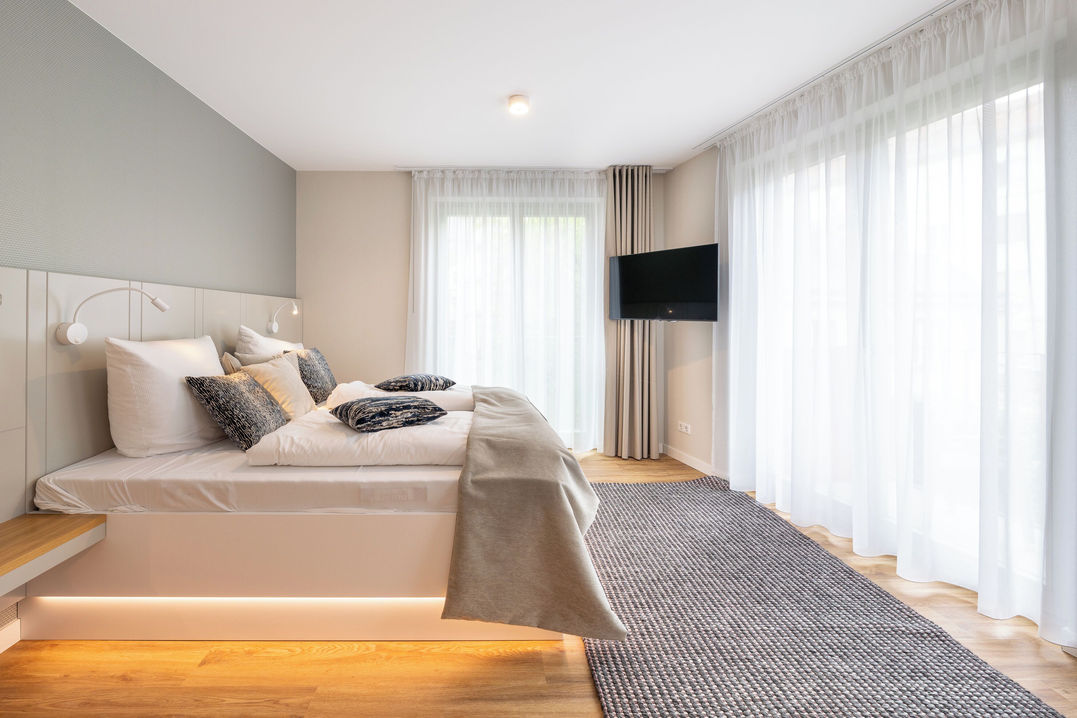A modern bedroom with a bed, bedside tables, a flat-screen TV, and sheer curtains.