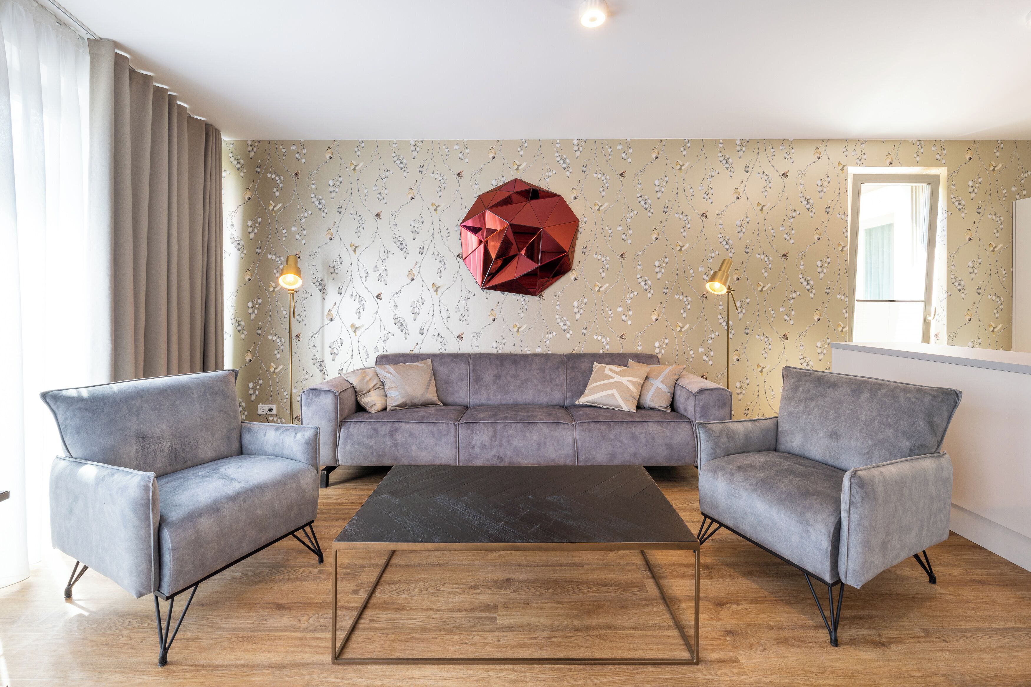En moderne stue med en grå sofa, to lænestole, et sort sofabord og en geometrisk vægdekoration.