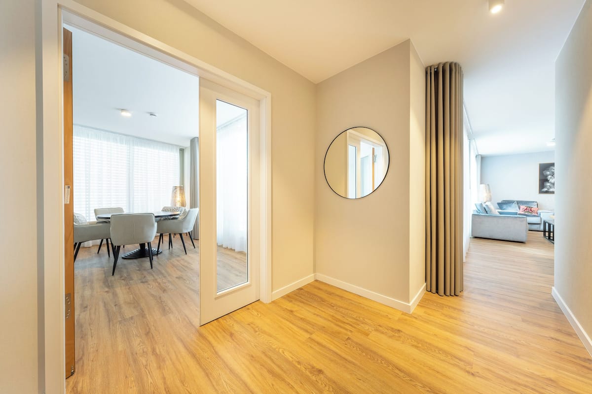A modern interior with a wooden floor, a dining area with chairs and a table, a living room with a sofa, and a large mirror on the wall.
