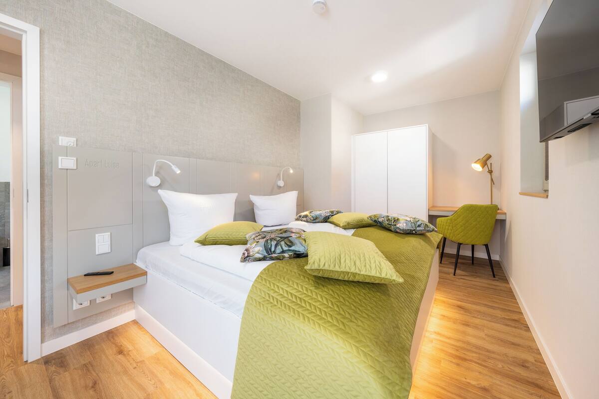A bedroom with a bed, bedside table, chair, and a TV.