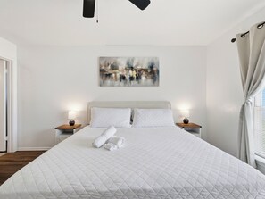 A neatly made bed with a quilted white duvet, two pillows, and rolled towels on a wooden nightstand. A painting is hung on the wall above the bed.