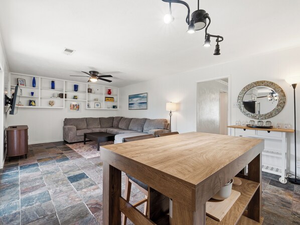 A living room with a sectional sofa, a wooden dining table, a ceiling fan, and a wall-mounted shelf with decorative items.