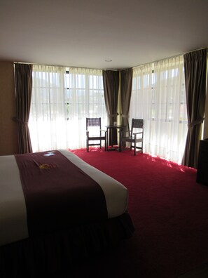 Ein Hotelzimmer mit einem großen Bett, zwei Stühlen, einem kleinen Tisch und durchsichtigen Vorhängen.