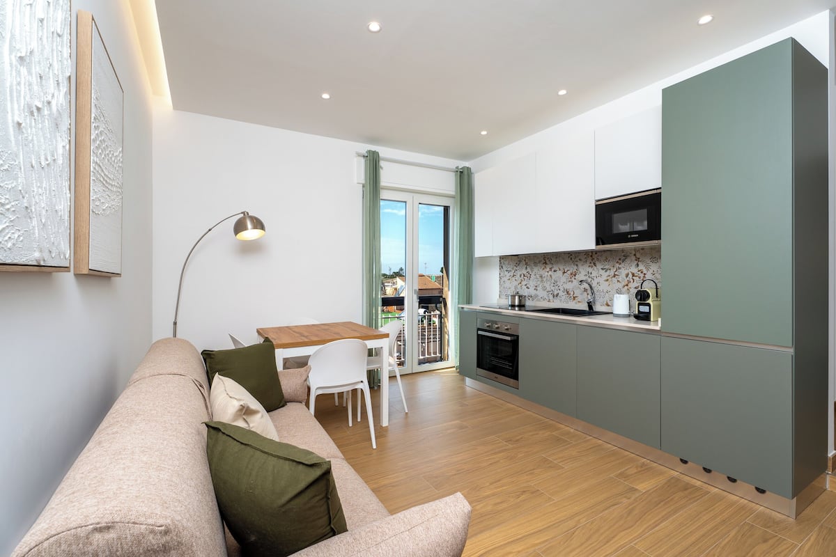 A modern kitchen with a dining area, a sofa with cushions, and a view through a glass door.
