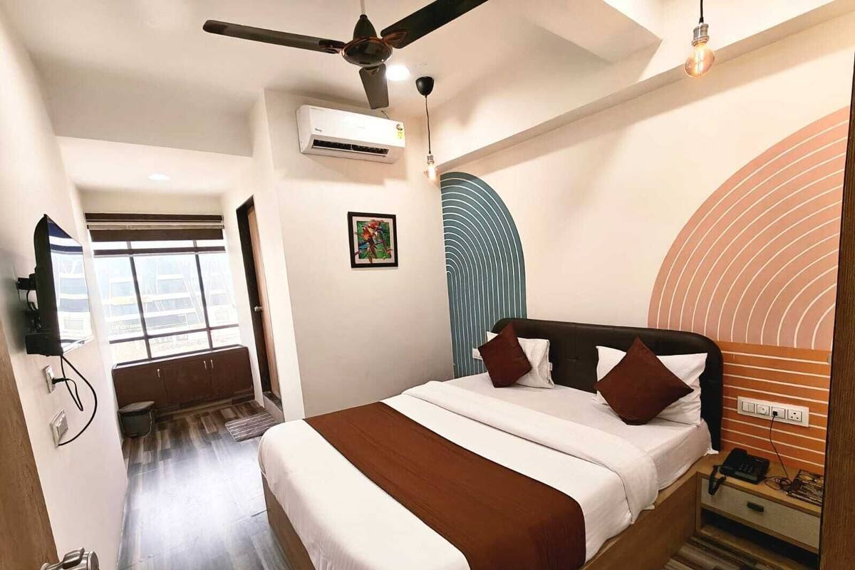 A hotel room with a bed, a ceiling fan, an air conditioning unit, a wall-mounted TV, and a window.