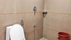 A bathroom with a toilet, shower, and a red bucket. - Hotel Krishna Inn (Ahmedabad)