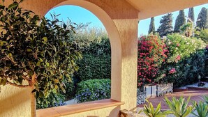 Un patio cubierto con un árbol en maceta, piso de baldosas y vistas a un jardín con diversas plantas y un muro de piedra.