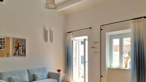 A living room with a sofa, a coffee table, a painting, a door, and a window with curtains. - YourHome - La Maddalena Blue House (La Maddalena)