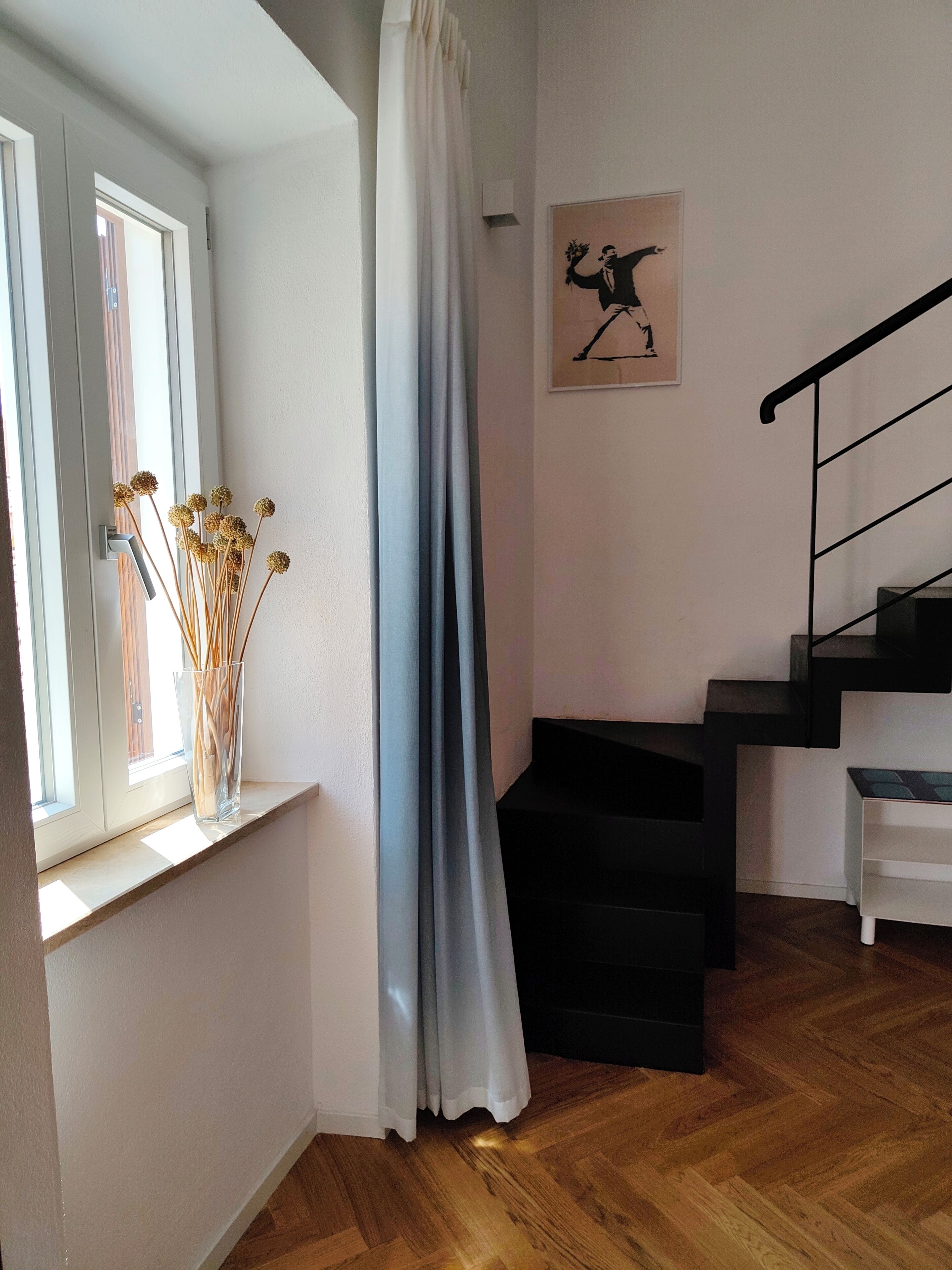 Une pièce avec une fenêtre, un rideau, un tableau encadré, un escalier et un plancher en bois.