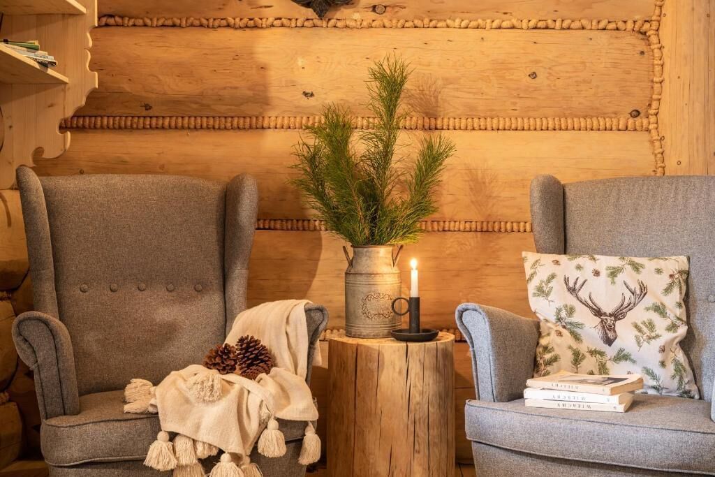 A cozy room with two grey armchairs, a wooden wall, a small wooden table with a candle, a pine branch, and a deer-patterned pillow.