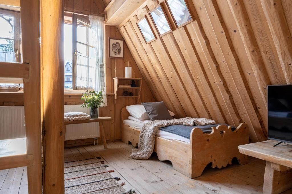A wooden room with a sloped ceiling, a bed, a small table, and a window with curtains.