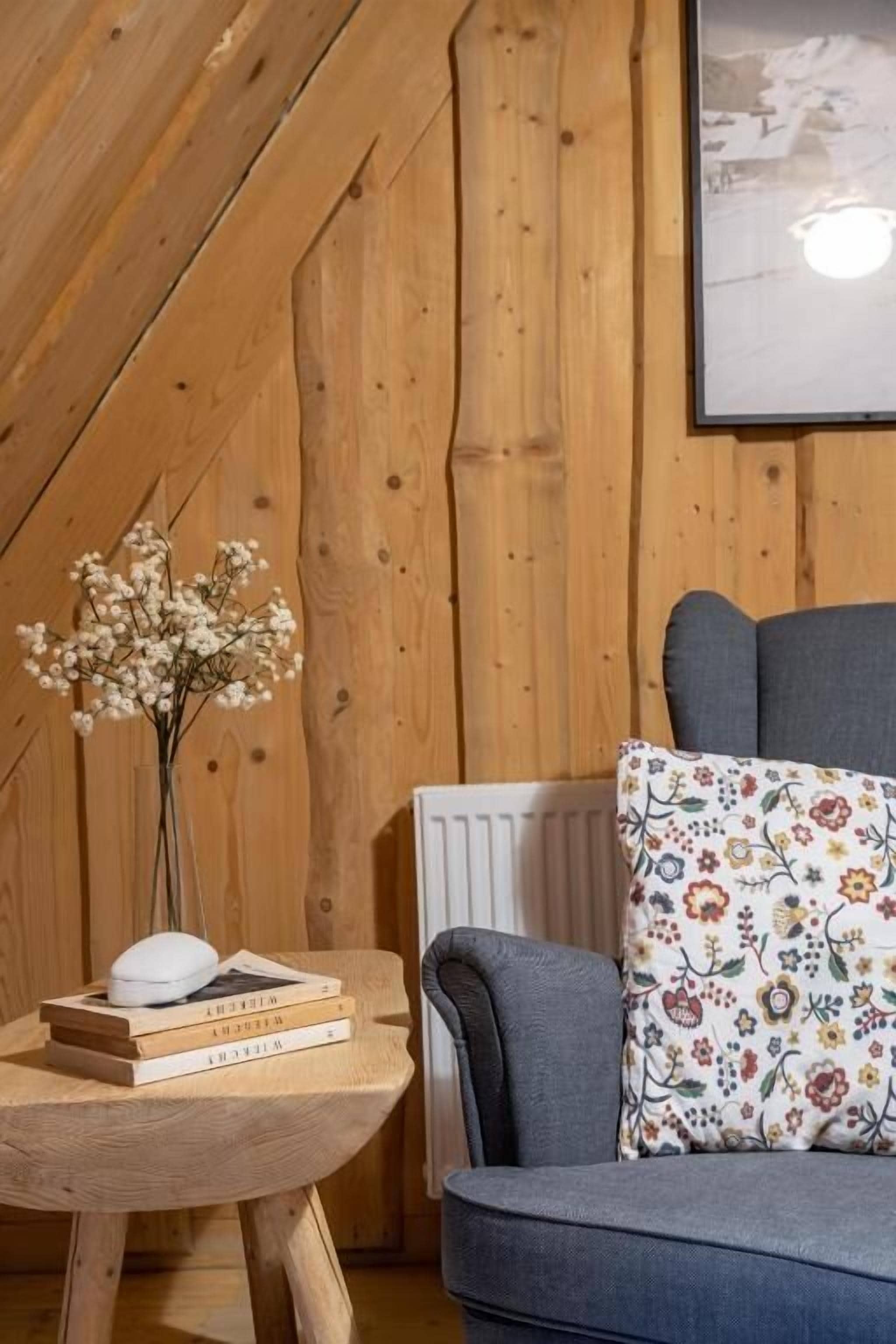Una habitación acogedora con una pared de madera, un sofá gris, un cojín floral, una pequeña mesa de madera con libros y un florero con flores.