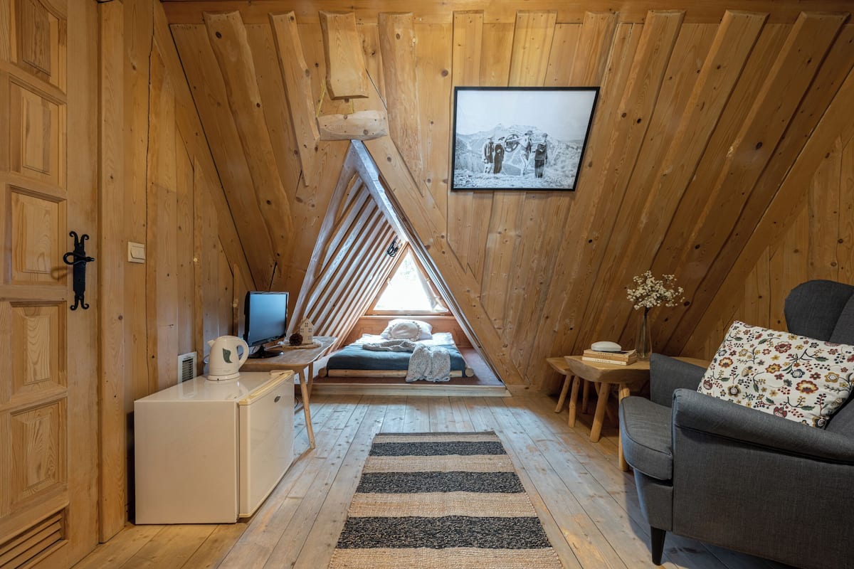 Un dormitorio en el ático con techo inclinado, una cama, un televisor, una mesita, una silla y un cuadro en la pared.