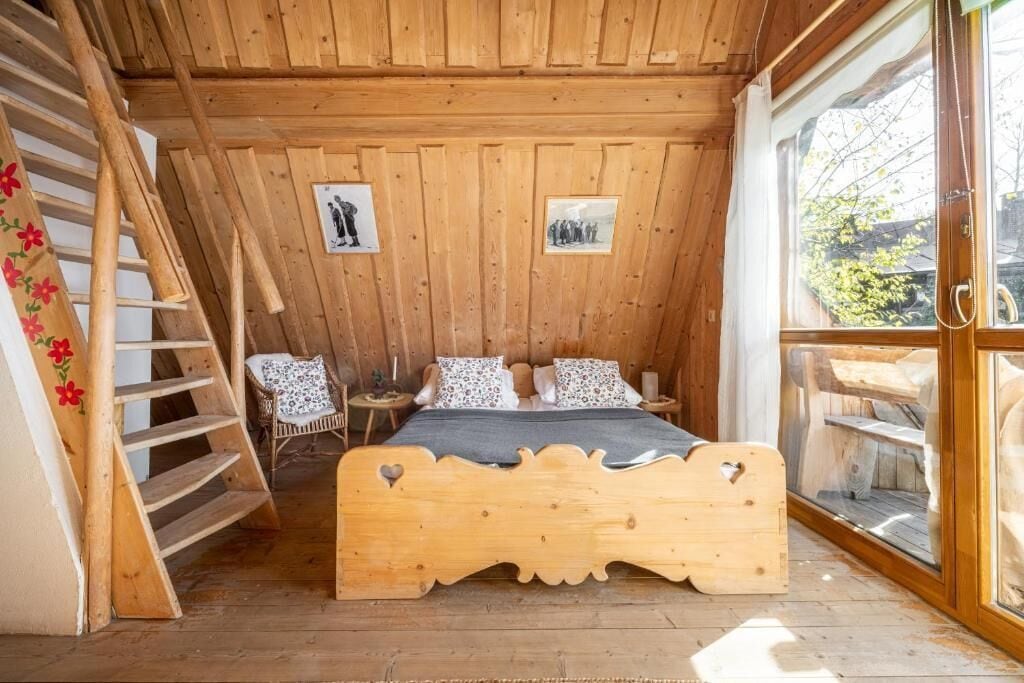 Un dormitorio de cabaña de madera con una cama, una silla, una escalera y un ventanal.