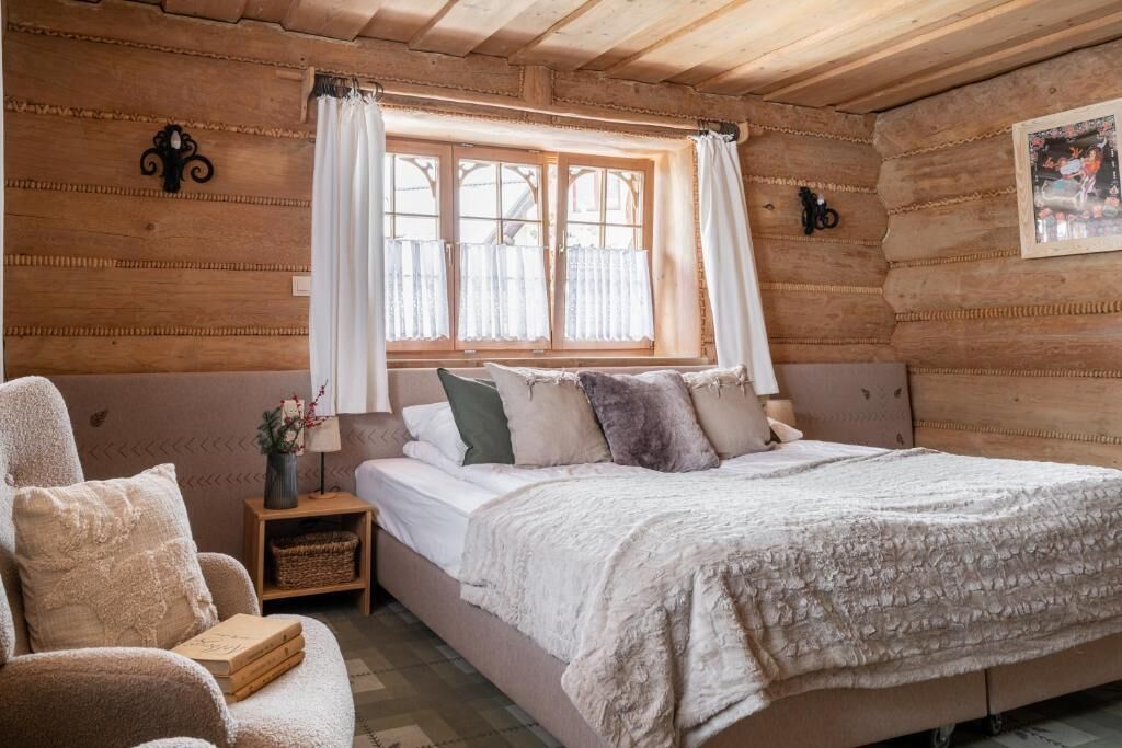 A cozy log cabin bedroom with a large bed, a chair, a nightstand, and a window with curtains.