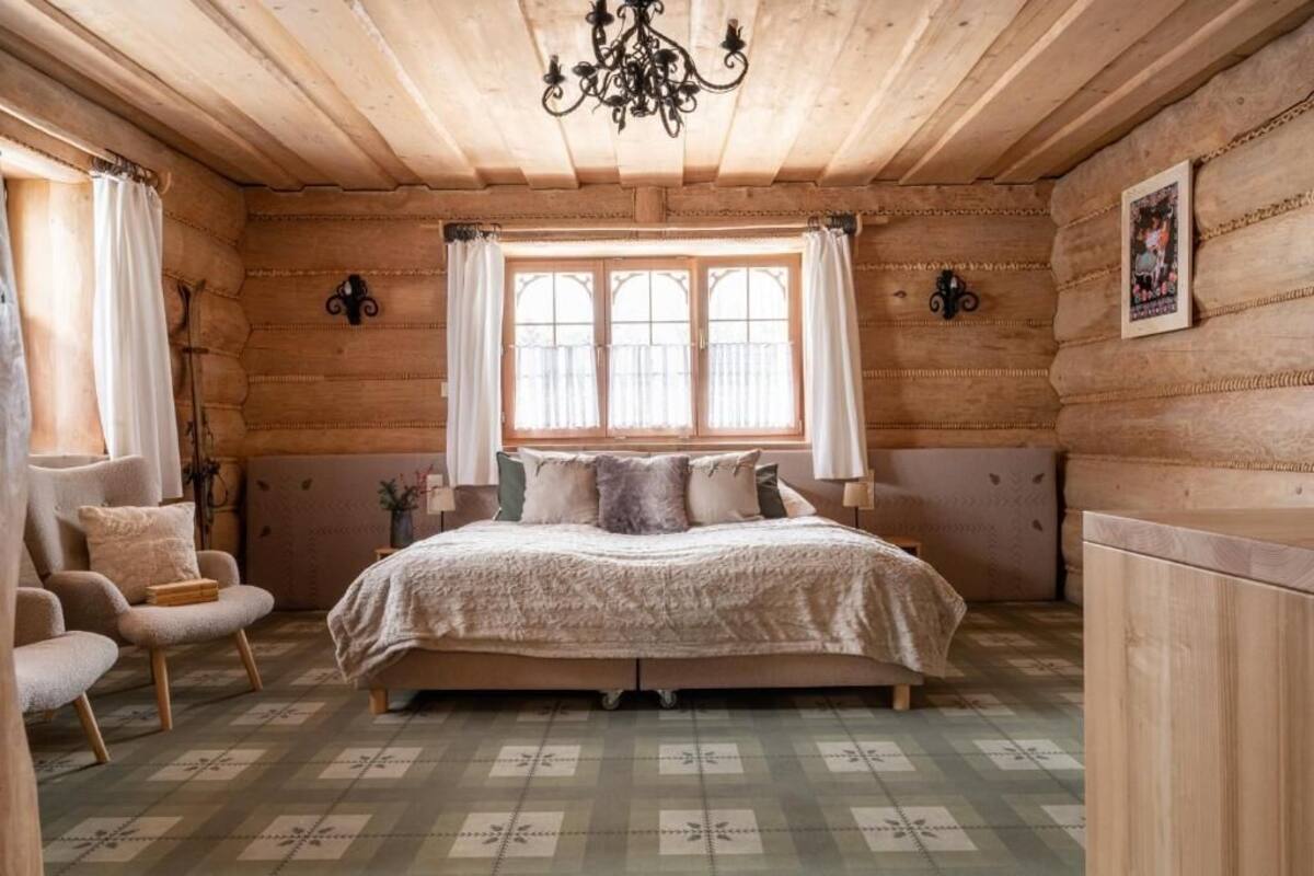Un dormitorio tipo cabaña de madera con una cama, dos sillas, una araña y un ventanal con cortinas.