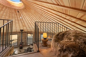 Ein Holzinnenraum mit hoher Decke, Kamin, rundem Tisch und einem mit Fell bezogenen Hocker.