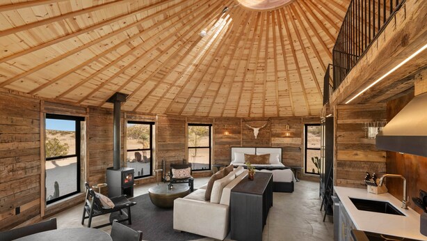 A spacious interior with a high wooden ceiling, a central fireplace, a sofa, a dining area, and a kitchen. - Mojave Yurt Featured in Rolling Stone, Stargaze (San Bernardino County)