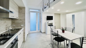 Een moderne keuken met een fornuis, spoelbak en kasten, een eethoek met een tafel en stoelen, en een tv bevestigd boven het aanrecht.