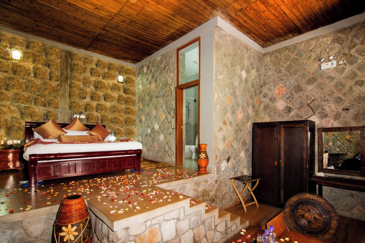 A bedroom with a stone wall, a wooden ceiling, a bed with pillows, a wooden cabinet, and a mirror.