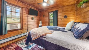 A wooden-paneled room with a bed, a ceiling fan, a mounted TV, and a window with curtains. - New! Hot Tub, Sauna, Arcade, cozy cottage - walk to everything! Great location! (Fayetteville)
