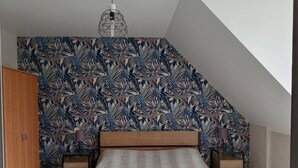 Un dormitorio con techo inclinado, una cama con colcha blanca, mesitas de noche de madera y una pared con motivos de hojas tropicales.