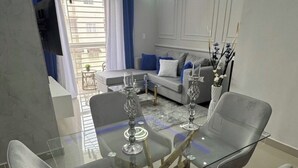 A dining area with a glass table and four chairs, a ceiling fan, and a living room in the background.