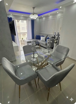 A dining area with a glass table and four chairs, a ceiling fan, and a living room in the background.