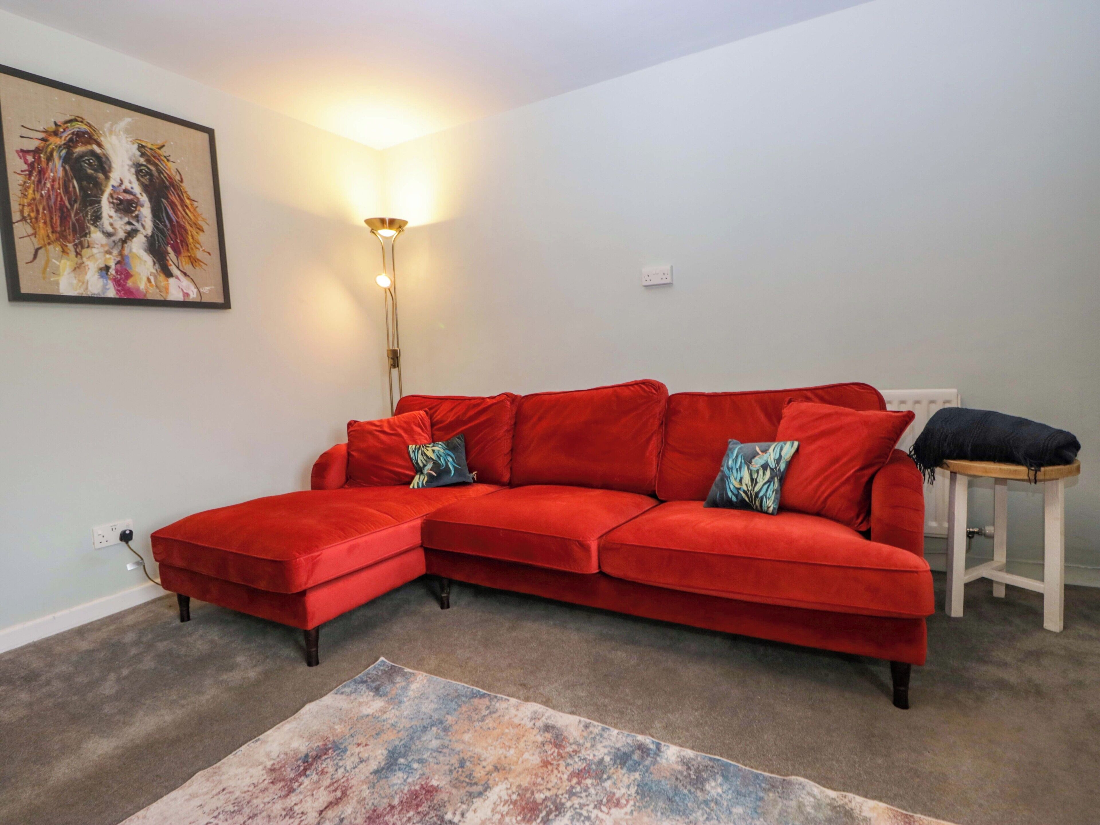 Ein rotes Ecksofa mit passendem Hocker, eine Stehlampe, ein farbenfroher Teppich und ein Bild an der Wand.