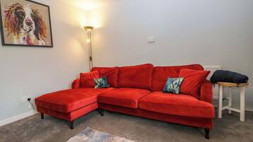 Ein rotes Ecksofa mit passendem Hocker, eine Stehlampe, ein farbenfroher Teppich und ein Bild an der Wand.