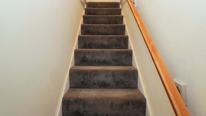 A carpeted staircase with a wooden handrail, white walls, and a white door at the top. - Dakota (Holmrook)