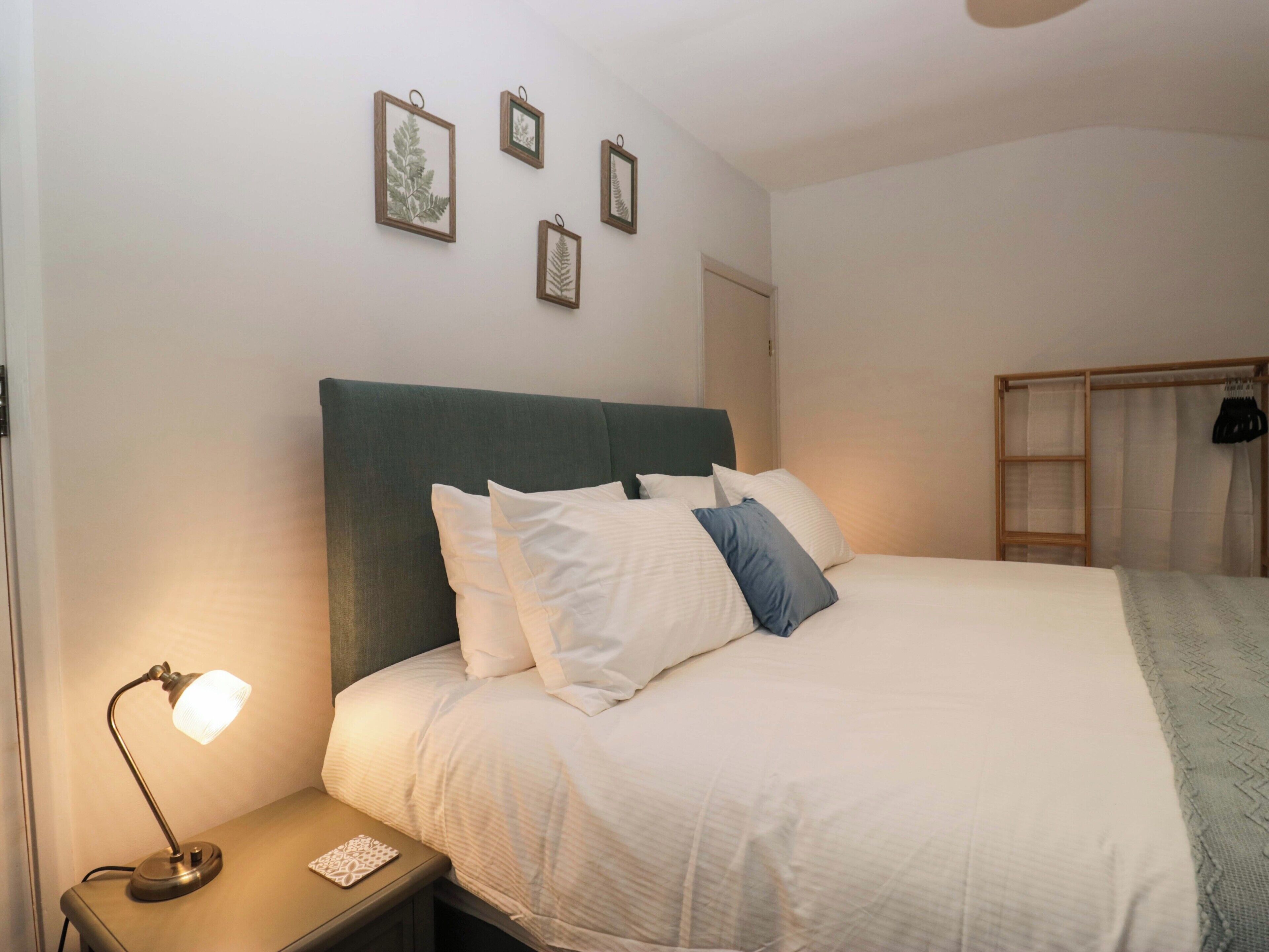Una cama bien tendida con almohadas blancas y azules, un cabecero verde, una lámpara de noche e impresiones botánicas enmarcadas en la pared.