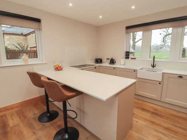 A modern kitchen with a central island, two bar stools, a built-in oven, and a window with a view of the outdoors. - The Gate House Annexe (Cockermouth)