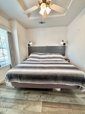 Una camera da letto con un letto, una ventola a soffitto, due abat-jour, una finestra con tende e un pavimento in legno.