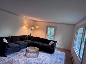 Un salon avec un canapé d’angle, une table basse ronde et un tapis à motifs. On y trouve des lampadaires et une fenêtre donnant sur un beau paysage.