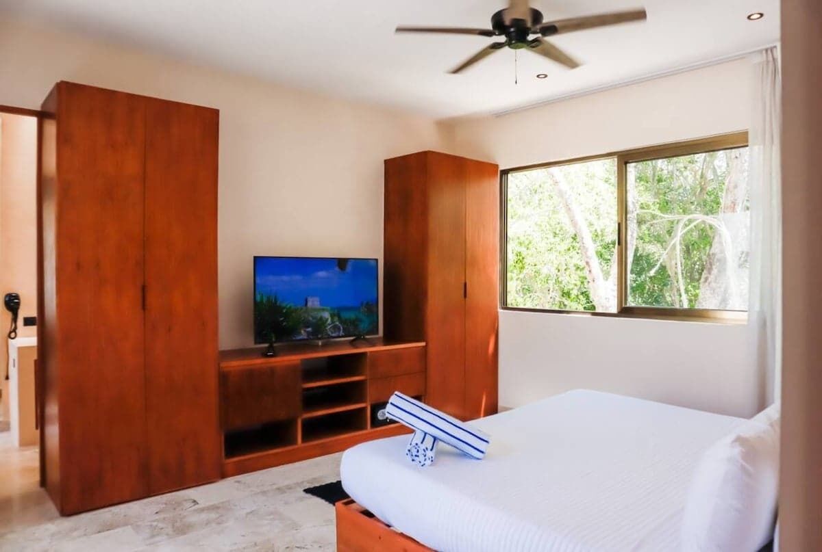 Ein Schlafzimmer mit einem Bett, einem Fernseher auf einem Holzständer, einem Deckenventilator und einem Fenster mit Blick auf Bäume.