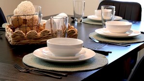 Une table à manger dressée avec des bols, des assiettes et des couverts blancs, accompagnée d’un panier de boules décoratives et d’un verre.