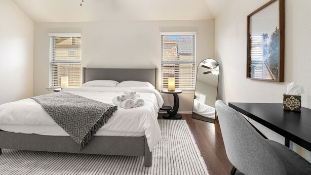 Une chambre à coucher comprenant un lit, des tables de chevet, un bureau, une chaise, un ventilateur de plafond et une fenêtre avec des stores.