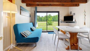 A room with a blue sofa, a wooden table, white chairs, a TV, and a sliding glass door to a garden. - Cosy Rural Cottage "Bergerie Couyrasseau" 1-5 Guests - Carcans - Wifi - Vacation/Remote Work (Carcans)