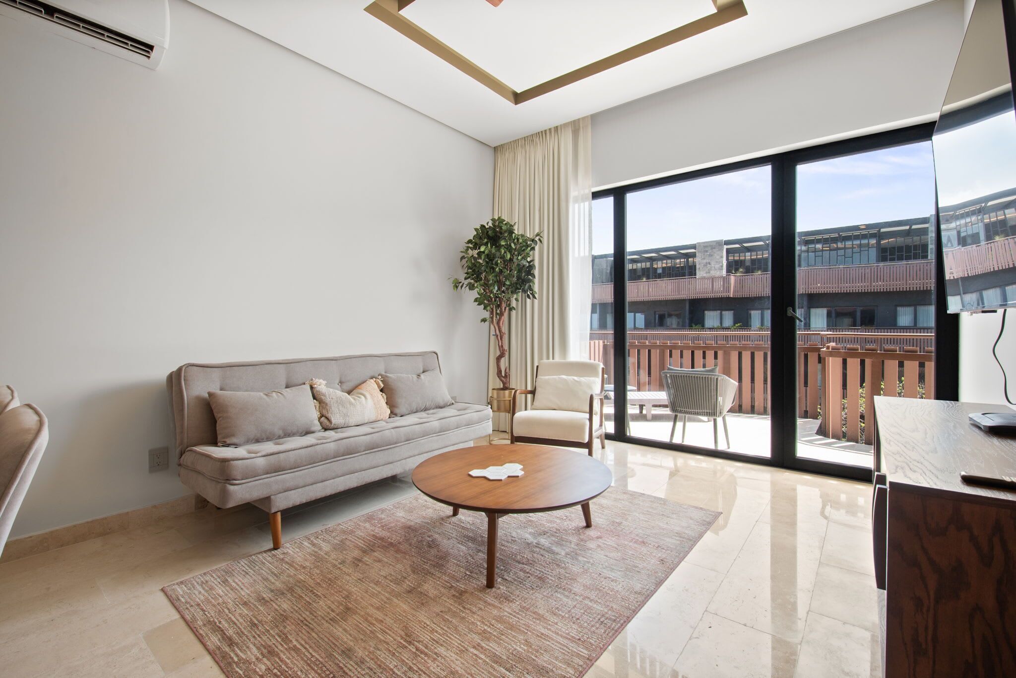 Un salon moderne comprenant un canapé, une table basse, un fauteuil, une plante et une grande fenêtre donnant sur un immeuble.